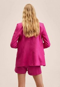 Personne aux longs cheveux blonds et ondulés, portant un blazer rose vif et un short assorti, debout devant un fond beige uni.