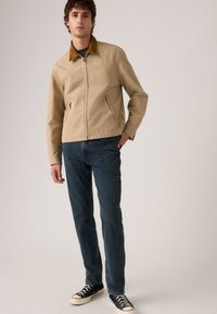 Μπεζ βαμβακερό μπουφάν με καφέ γιακά από κοτλέ, διαθέτει πλαϊνές τσέπες και κλείσιμο με φερμουάρ. Φορεμένο με σκούρο μπλε τζιν και μαύρα αθλητικά παπούτσια.