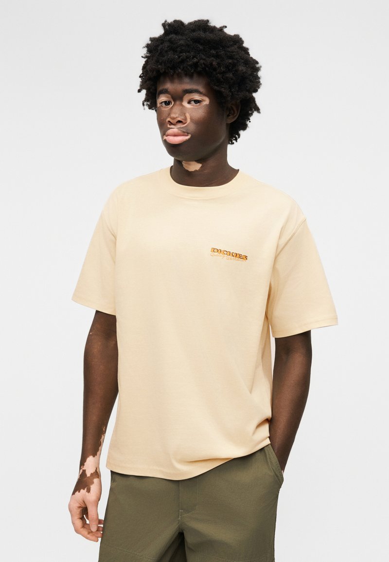 Junger Mann mit Vitiligo, trägt ein beiges Dickies-T-Shirt und olivgrüne Hosen, steht mit einer Hand in der Tasche vor einem schlichten Hintergrund.