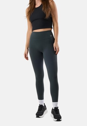 Frau trägt schwarzen Crop-Top, hoch taillierte dunkelgraue Leggings mit Seitentasche, weiße Socken und schwarze Laufschuhe, steht vor weißem Hintergrund.