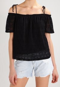 Haut noir épaules dénudées avec détails en dentelle, encolure élastique et manches courtes. Associé à un short en jean clair avec ourlet effiloché.