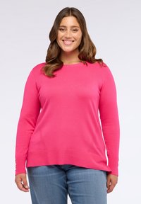 Donna sorridente con capelli castani, indossa un maglione a maniche lunghe rosa acceso e jeans blu, in piedi contro uno sfondo bianco semplice.