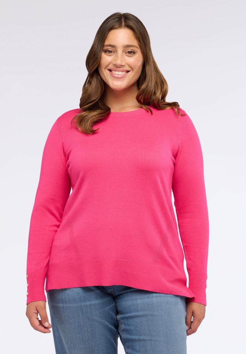 Donna sorridente con capelli castani, indossa un maglione a maniche lunghe rosa acceso e jeans blu, in piedi contro uno sfondo bianco semplice.