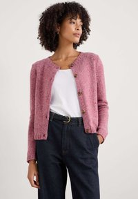 Różowy dziewiarski kardigan z okrągłym dekoltem, zapi�ęciem na guziki i ściągaczami przy mankietach. Nosi się go na białej koszuli z ciemnoniebieskimi spodniami o wysokim stanie.