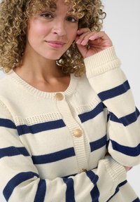 Gilet crème avec des rayures horizontales bleu marine. Doté d'un col rond, d'une fermeture à boutons et de poignets côtelés, fabriqué en tissu tricoté doux.