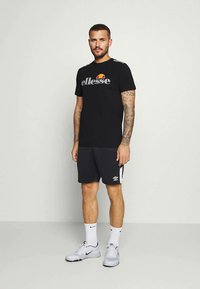 Black short-sleeve T-shirt with "ellesse" logo and multicolored accent. Paired with dark shorts and white sneakers. Simple athletic design.