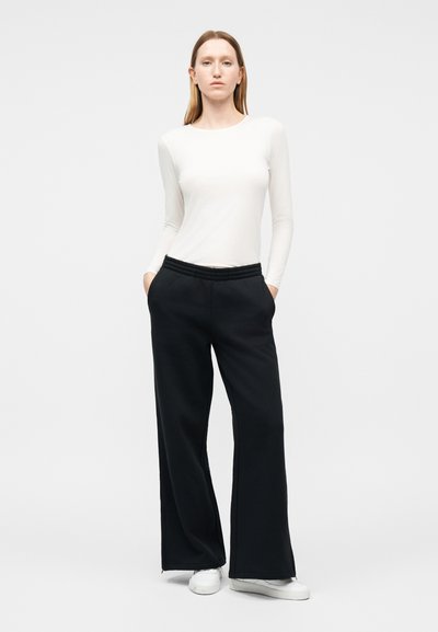 Γυναίκα που στέκεται με τα χέρια στις τσέπες ενός μαύρου παντελονιού με φαρδιά μπατζάκια, φοράει λευκή μπλούζα με μακριά μανίκια και λευκά αθλητικά παπούτσια σε απλό λευκό φόντο.