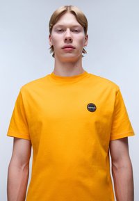 Camiseta de algodón de manga corta y cuello redondo en color naranja brillante, con un parche de logo negro circular en el lado izquierdo del pecho.
