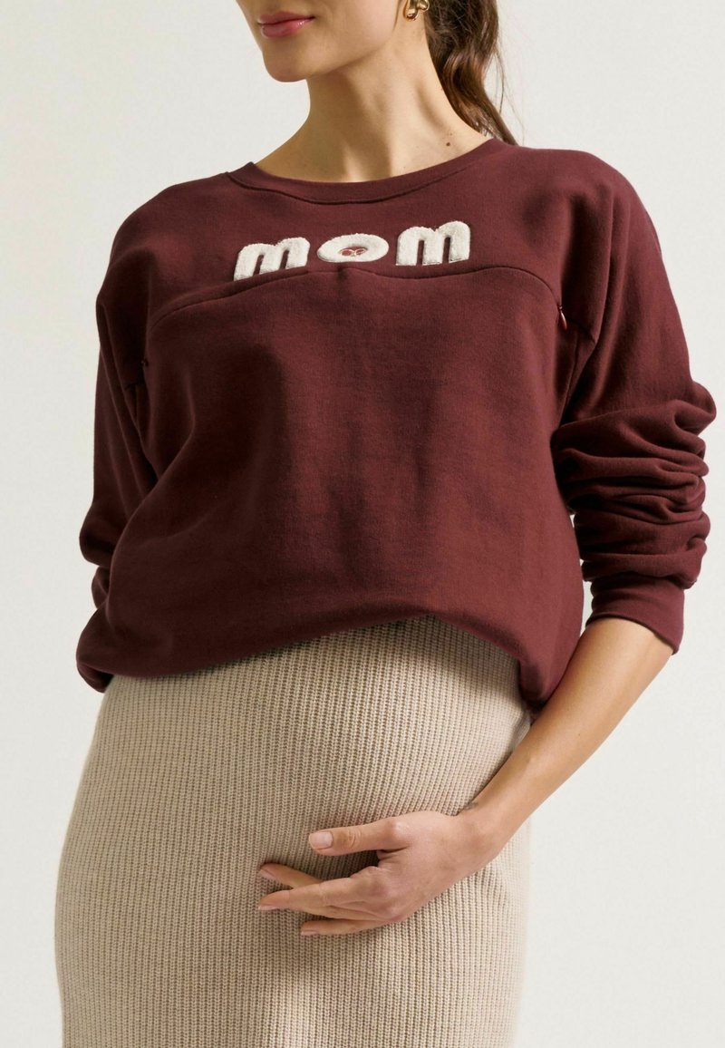 Bordeaux sweatshirt met geborduurd "mom", losse pasvorm, geribde manchetten, gedragen over een gestructureerde beige gebreide jurk die de taille bedekt.