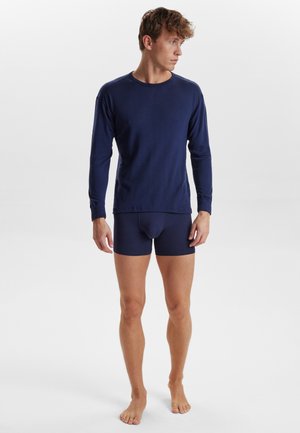 Langærmet navy top i blødt stof parret med navy boxer shorts, der har et tætsiddende design og glat tekstur. Modellen står barfodet.