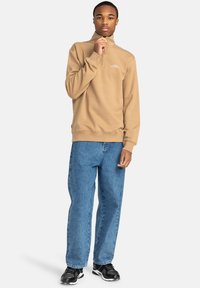 Felpa zipata color sabbia con colletto alto, abbinata a jeans in denim blu larghi. Il modello indossa scarpe sportive nere, mostrando un look casual.