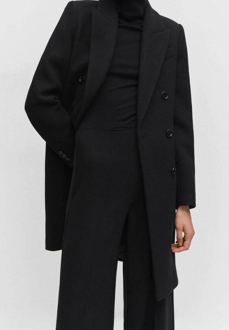 Sort dobbeltbrystet frakke med store reverser, lavet af et struktureret stof. Har knapper, lommer og en skræddersyet pasform over en sort top og bukser.