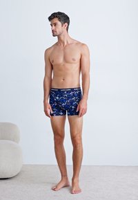 Blue patterned boxer shorts with a black waistband, displaying a mix of shades and abstract shapes. Model stands barefoot against a neutral background.
