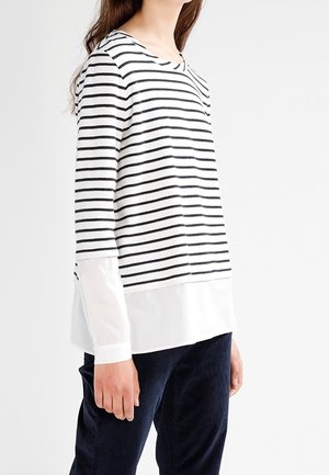 Mujer con un top de manga larga de rayas horizontales en blanco y negro con dobladillo blanco en capas y pantalones azul marino, de pie contra un fondo blanco.