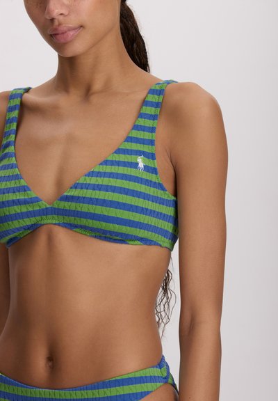 Γυναίκα που φοράει μπικίνι με πράσινες και μπλε ρίγες και λευκό κεντημένο λογότυπο στη αριστερή πλευρά, με φόντο απλό.