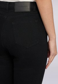 Jeans de mezclilla negra con una cintura alta, una superficie texturizada y un bolsillo trasero con un diseño puntiagudo. La cintura tiene una etiqueta de cuero.
