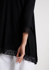 Kvinnas hand vilar vid sidan, iförd en svart långärmad topp med spetsdetaljer på ärmar och fåll, tillsammans med vita byxor.