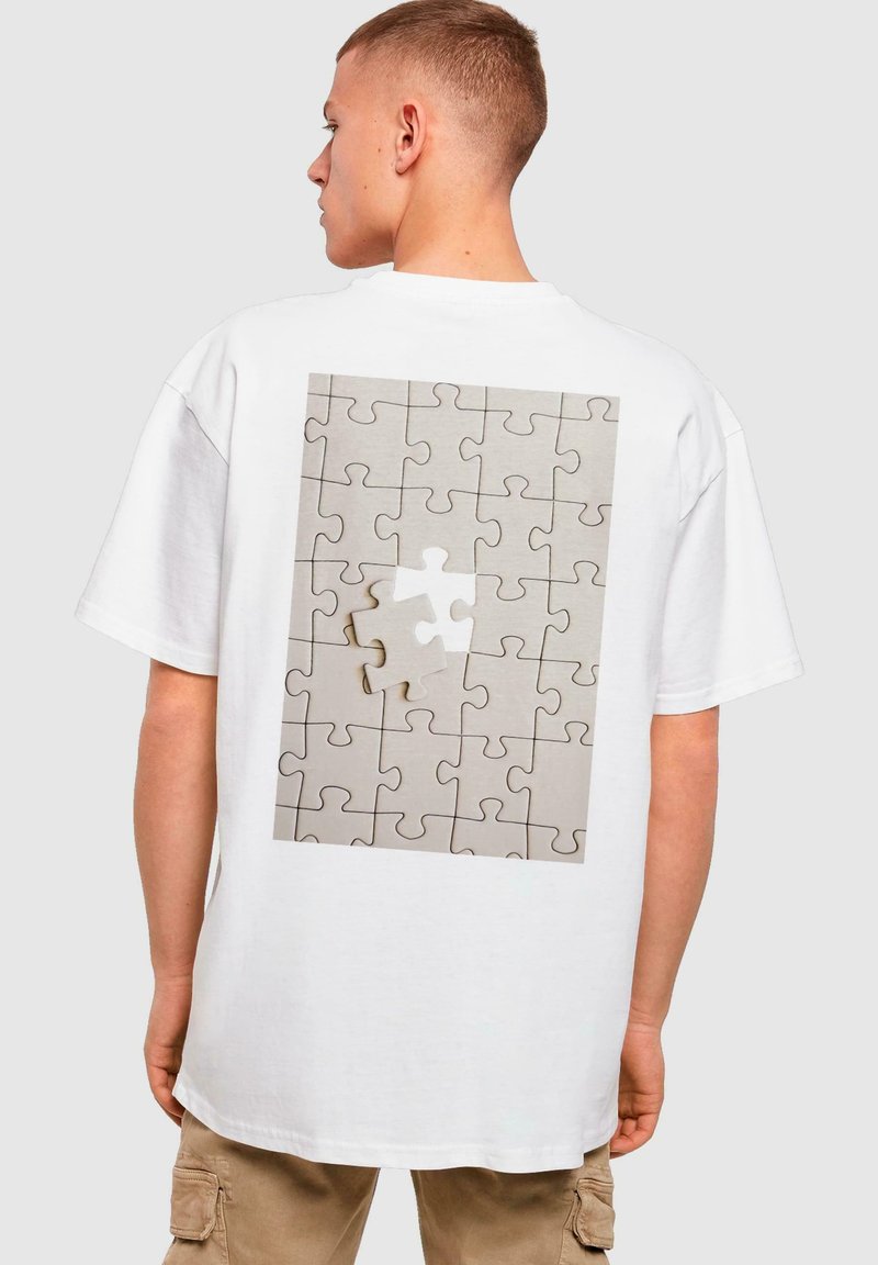 Hvid oversized t-shirt med et trykt design af et lysegråt puslespil med én brik, der mangler, hvilket skaber kontrast.