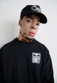 Fekete pulóver fehér "OBEY" logóval a mellrészen és fekete sapka hímzett logóval. Simma szövet és laza szabás.