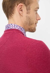 Roter Strickpullover mit rundem Ausschnitt, strukturierter Stoff; darunter ein Hemdkragen mit floralem Muster in Blau, Pink und Weiß.