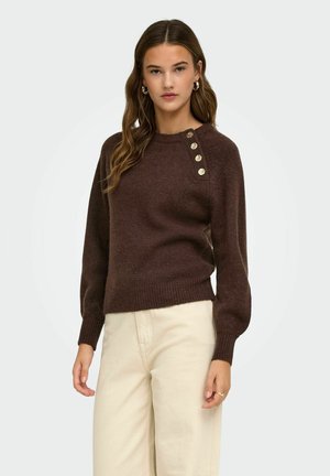 Mujer que lleva un suéter marrón oscuro con botones dorados en el hombro y pantalones beige de cintura alta, de pie frente a un fondo liso.