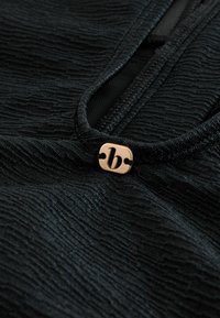 Zbliżenie na teksturowaną czarną tkaninę z małą metalową metką z stylizowaną małą literą "b" przy dekolcie ubrania.