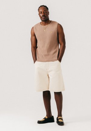 Mies seisoo kädet taskuissa, yllään hihaton beige ribbineuletoppi, kermaan vivahtavat polvipituuiset shortsit, mustat ja vaaleanruskeat loaferit sekä kultaiset kaulakorut.