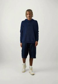 Felpa blu marina con tasca frontale e polsini a maglia coordinati, abbinata a pantaloni corti in denim fino al ginocchio con un piccolo logo, e sneakers bianche.