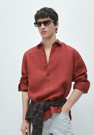 Junger Mann mit einem rostfarbenen Hemd mit Knöpfen, Sonnenbrille, hellen Jeans und einem dunkel karierten Hemd, das um seine Taille gebunden ist.