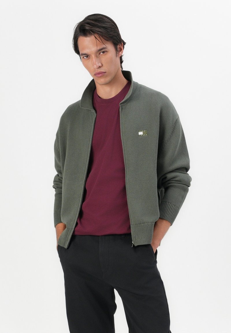 Khaki zip-up cardigan with ribbed cuffs and collar; worn over a burgundy shirt; features a small logo detail on the chest.