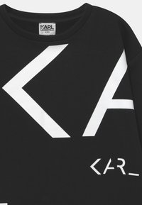 Černá mikina s velkými bílými geometrickými tvary a textovým designem. Materiál je měkký, s kulatým výstřihem a žebrovanými manžetami.