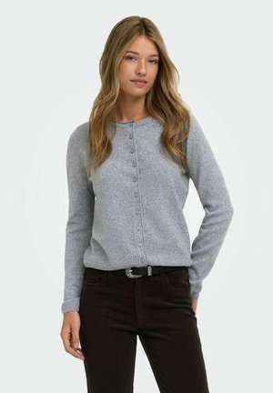 Femme aux longs cheveux blonds portant un cardigan gris clair boutonné et un pantalon foncé avec une ceinture noire, debout devant un fond uni.