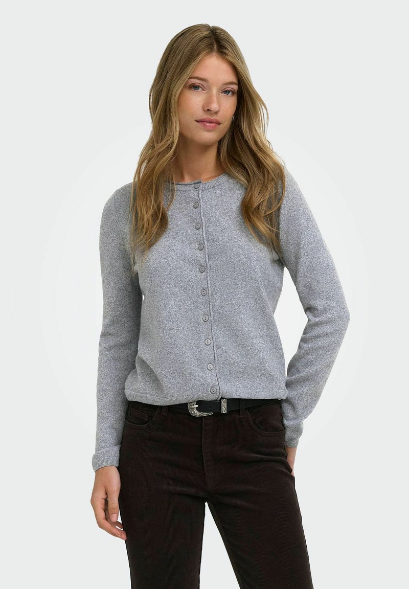 Femme aux longs cheveux blonds portant un cardigan gris clair boutonné et un pantalon foncé avec une ceinture noire, debout devant un fond uni.