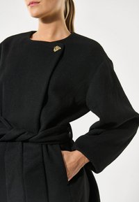 Juodas vilnonis paltas su struktūrizuotu dizainu, turintis apvalų iškirpimą, ilgomis rankovėmis, diržo juosmeniu ir auksine detalė prie apykaklės.