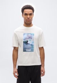 Άνδρας που φορά λευκό T-shirt με γραφικό κύματος ωκεανού και το κείμενο «HUGO», συνδυασμένο με μαύρο παντελόνι, στέκεται μπροστά σε απλό ανοιχτό φόντο.