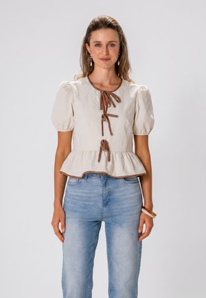 Frau trägt eine beige Bluse mit Puffärmeln und braunen Schleifenbändern sowie hellblaue, hoch taillierte Jeans und steht vor einem schlichten Hintergrund.