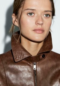 Veste en cuir marron avec grand col, dotée d'une fermeture éclair et d'accents en bouton noir. Texture lisse avec un léger éclat.