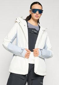 Weißer und hellblauer Skijacke mit einer gefütterten Kapuze, Reißverschluss und elastischen Bündchen, getragen über einem dunklen Oberteil, mit einer Sonnenbrille.