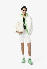 Veste en denim blanc sur un hoodie vert, chemise à fleurs, shorts blancs et chaussures de sport vertes. Portant un sac fourre-tout léger. Lunettes de soleil portées.
