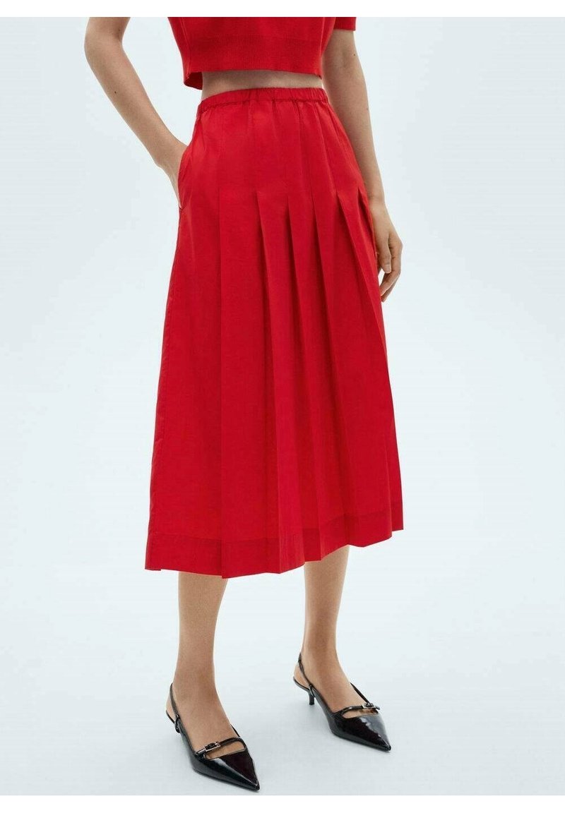 Femme portant une jupe midi plissée rouge taille haute avec poches, associée à des escarpins slingback noirs pointus sur fond blanc.