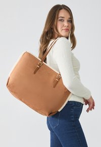 Sac fourre-tout beige avec une doublure en fausse fourrure crème douce, poignées doubles et accents en métal doré. Design simple, forme rectangulaire, texture robuste.