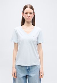 Γυναίκα με ίσια καστανά μαλλιά φοράει ανοιχτό μπλε μπλουζάκι με V στο λαιμό και άνετο μπλε τζιν, στέκεται μπροστά σε απλό ανοιχτό φόντο.