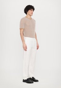 Chemise en tricot gris à manches courtes avec col marron, assortie à un pantalon en lin blanc et des chaussures en cuir noir. Le modèle se tient devant un fond blanc.