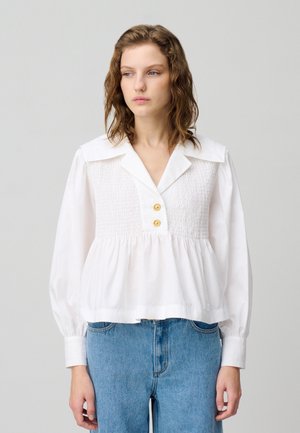 Blouse en coton blanc avec un décolleté en V profond, un large col et un corps froncé. Présente des manches bouffantes longues et deux boutons dorés à l'avant.