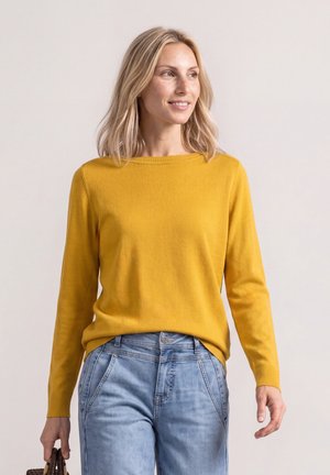 Lyshåret kvinde, der smiler, iført en sennepsgul sweater og lyseblå jeans, holder en lille brun håndtaske i venstre hånd.