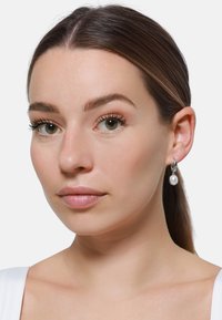 Orecchini a cerchio in argento con una perla pendente, caratterizzati da una superficie liscia e una finitura lucida, indossati da una modella con trucco neutro.