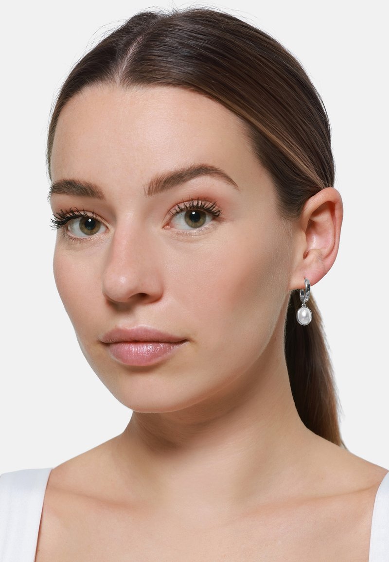 Orecchini a cerchio in argento con una perla pendente, caratterizzati da una superficie liscia e una finitura lucida, indossati da una modella con trucco neutro.