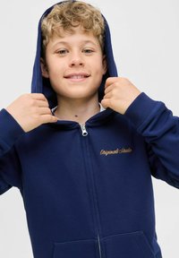 Marinblå zip-hoodie i mjukt tyg med en stor huva, med dragkedja fram och broderad guldtext "Originals Studio" på bröstet.