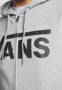 Sudadera con cremallera gris hecha de tela suave, que presenta un gran logo negro de "VANS". Incluye cordones ajustables y cremallera frontal.