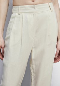 Pantalones de pierna ancha beige, hechos de tela texturizada, con plisados en la parte delantera y una cintura alta. Incluyen presillas para cinturón y bolsillos laterales.